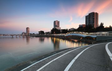 Paved pathway next to calm lagoon waters flowing underneath bridge with apartment buildings in backdrop against pastel sunset in Nanaimo BC Vancouver Island Canada